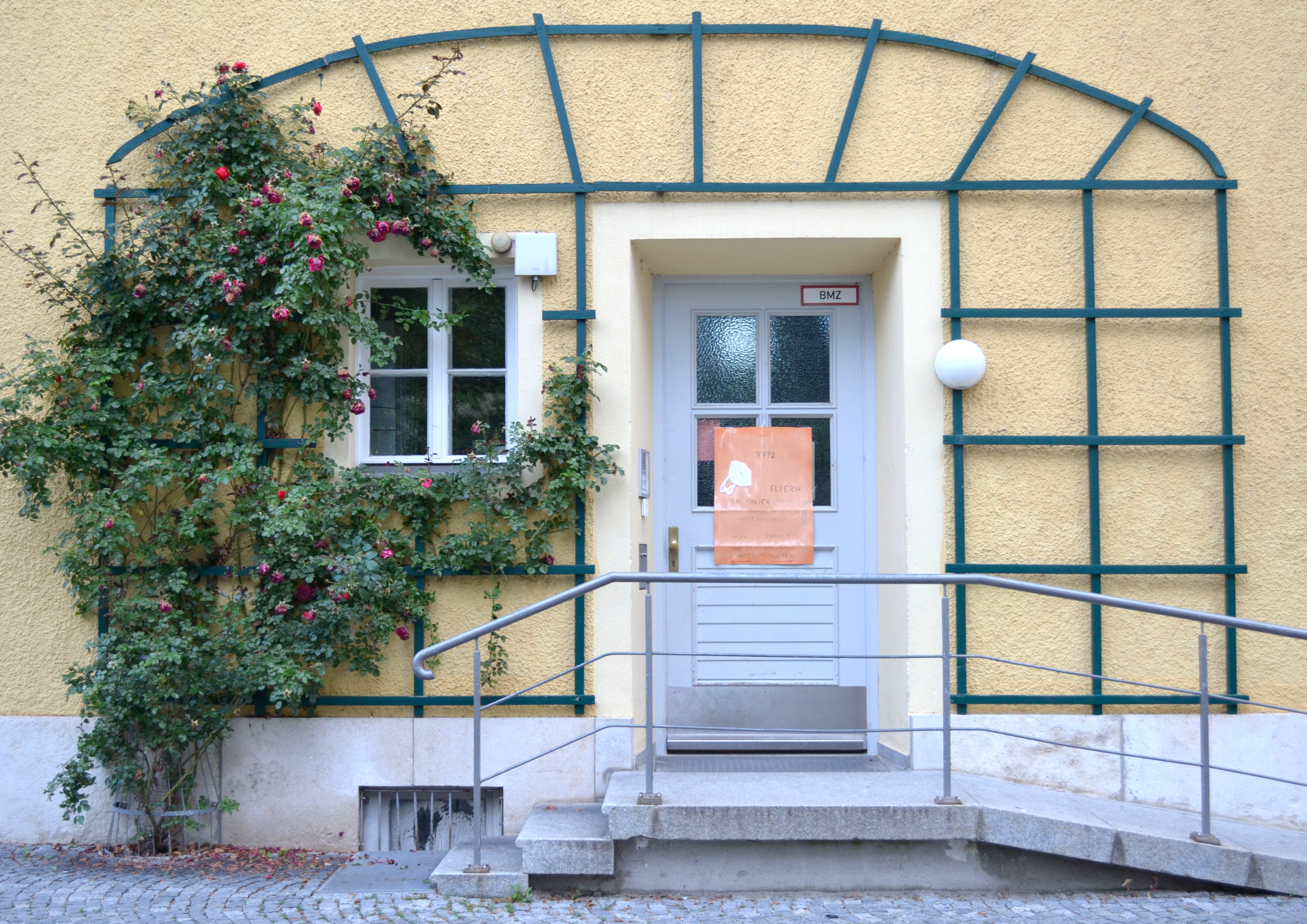 Vor dem Eingang eines gelben Hauses, das mit Efeu bewachsen ist, sind sowohl drei Stufen als auch gegenüber eine Rampe.